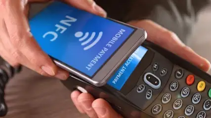 Dłonie trzymają telefon z NFC nad terminalem płatniczym. Na ekranie telefonu i terminalu widoczne informacje o płatności.