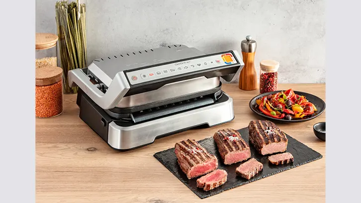 Na drewnianym stole stoi srebrny grill elektryczny, talerz sałatki warzywnej i deska z pokrojonym mięsem.