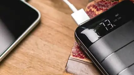 Na drewnianym stole leżą: smartfon, czarny powerbank z wyświetlaczem 73% i kabel.