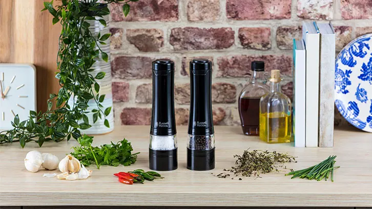 Na stole znajdują się przyprawy, zioła, czosnek, chili, młynki do soli i pieprzu oraz butelki z płynami. W tle widać ceglaną ścianę i książki.