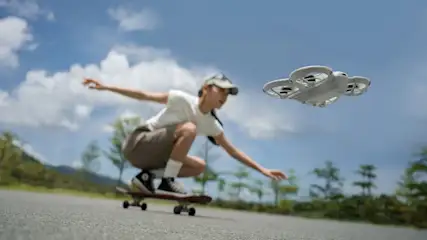 Kobieta na deskorolce, na tle nieba i zieleni. Nad nią unosi się biały dron.