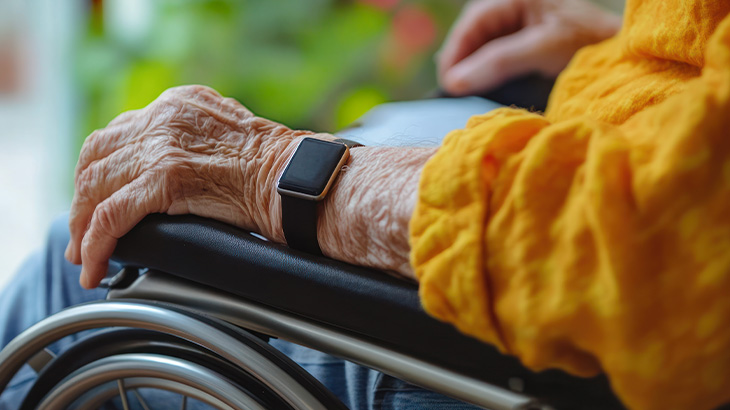 Dłoń osoby z zegarkiem spoczywa na wózku inwalidzkim, obok żółta tkanina, w tle zieleń.