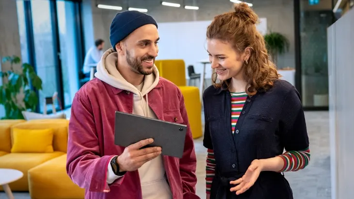 Mężczyzna w bordowej kurtce trzyma tablet, uśmiecha się do kobiety w ciemnej bluzce w paski. W tle widać żółte sofy i rośliny.