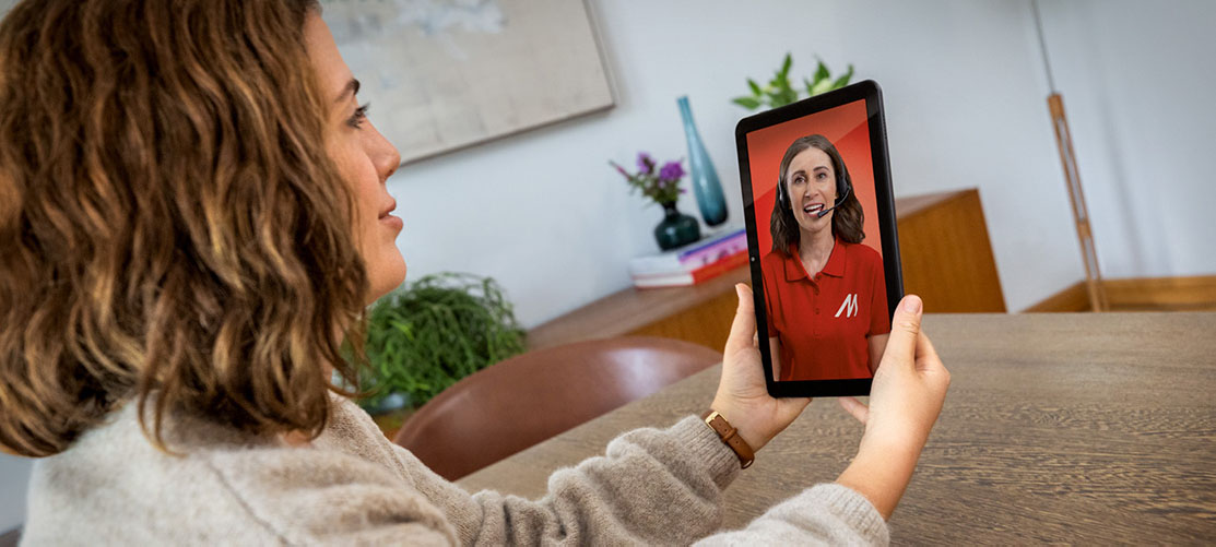 Kobieta trzyma tablet z wideorozmową z konsultantką w czerwonej koszulce z logo. W tle widać stół i dekoracje w pomieszczeniu.