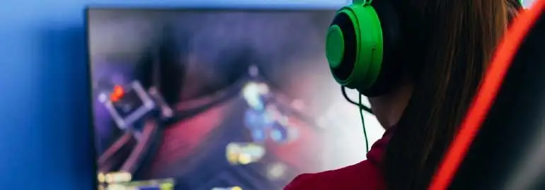Osoba w zielonych słuchawkach gra w grę na monitorze na tle niebieskiej ściany, siedząc na krześle z czerwonymi akcentami.