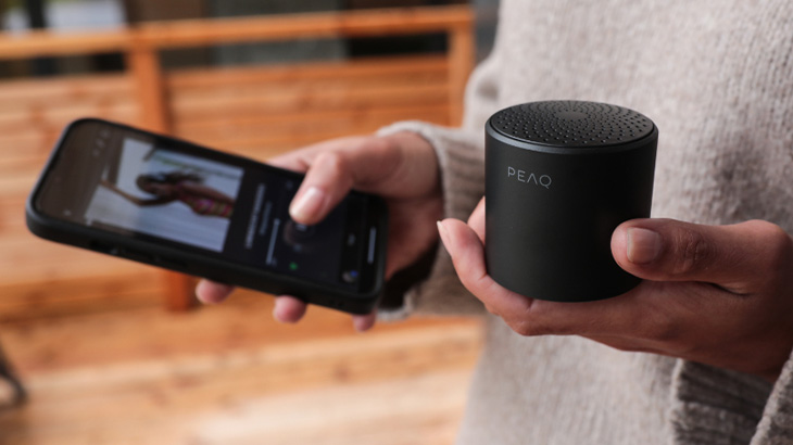 Osoba trzyma czarny telefon i głośnik na tle drewnianej balustrady.