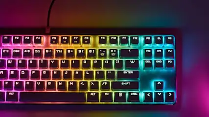 Klawiatura z podświetleniem RGB i mysz na fioletowym tle. Kabel klawiatury biegnie w górę kadru.