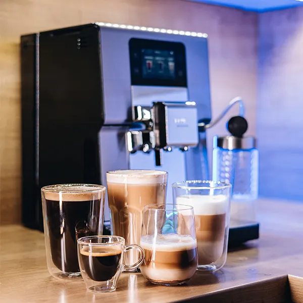 Na blacie stoi ekspres do kawy z różnymi rodzajami kaw w szklankach. Tło jest niebieskie.