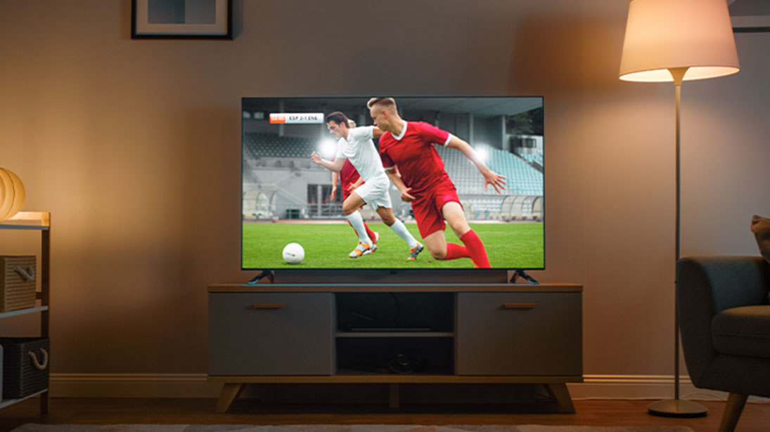 TV 65 cali na stoliku w salonie, z meczem piłkarskim na  ekranie. Obok włączona lampa. 