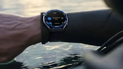 Osoba w czarnym kombinezonie ma na nadgarstku smartwatch z niebieską obwódką, w wodzie.