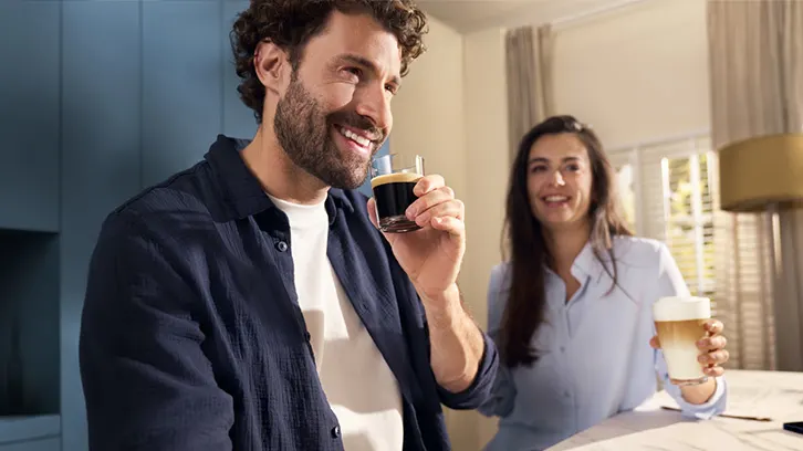 Mężczyzna trzyma szklankę kawy, uśmiecha się. Za nim kobieta siedzi przy stole z kawą. W tle okno i zasłony.