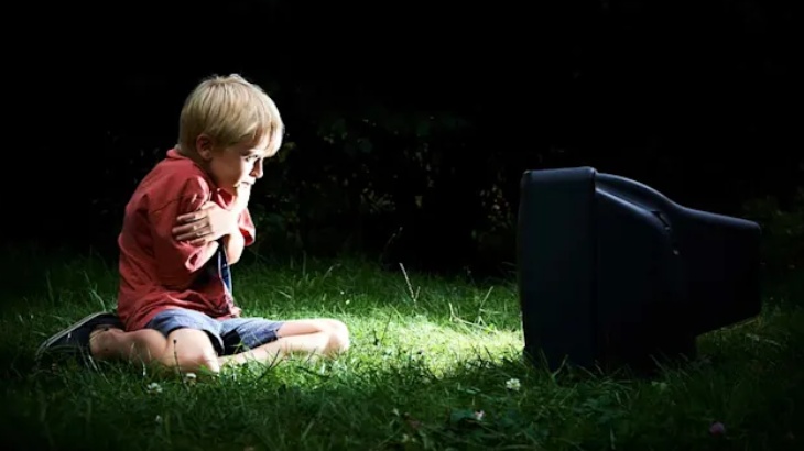 Chłopiec w czerwonej koszulce siedzi na trawie, obejmuje się ramionami i patrzy na czarny telewizor. Ciemne tło.