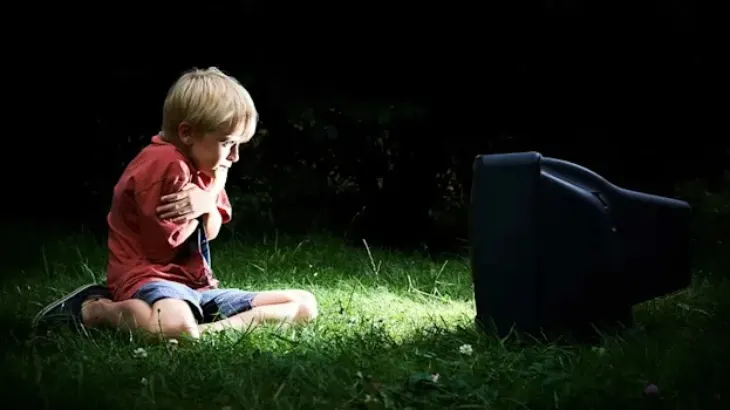 Chłopiec w czerwonej koszulce siedzi na trawie, obejmuje się ramionami i patrzy na czarny telewizor. Ciemne tło.