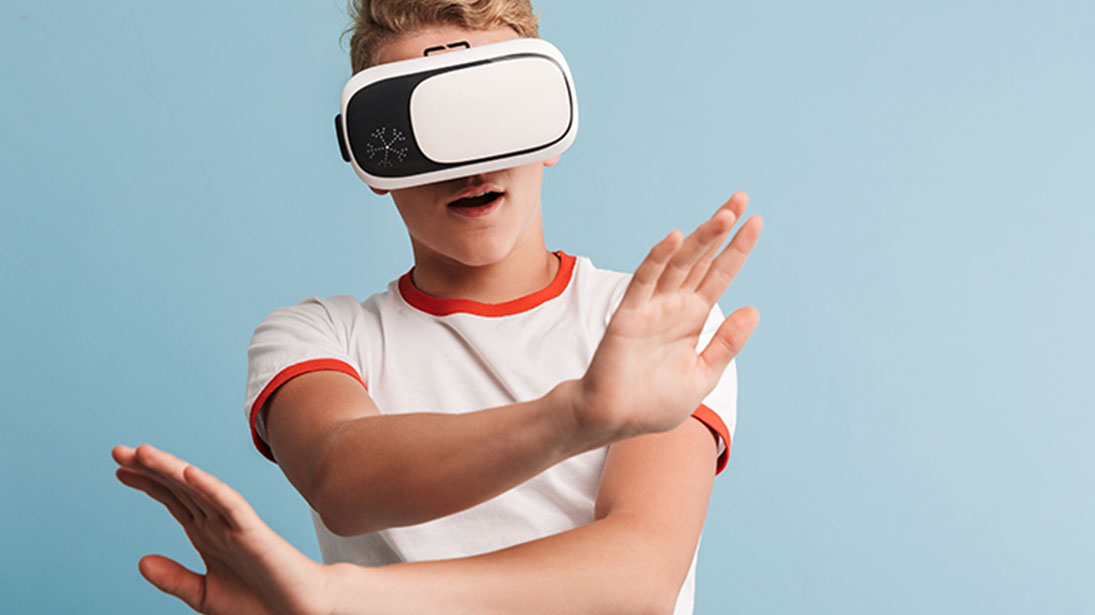 Chłopiec w białej koszulce z czerwonym wykończeniem używa białych gogli VR na tle niebieskiego tła, wyciągając ręce.