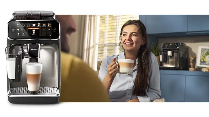 Kobieta pije kawę w kuchni. Ekspres do kawy Philips stoi obok, przygotowując napój. W tle widać okno i niebieskie szafki.