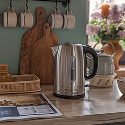 Na blacie kuchennym stoi czajnik ze stali nierdzewnej. W tle widoczne deski do krojenia, kwiaty w wazonie i wiszące kubki.