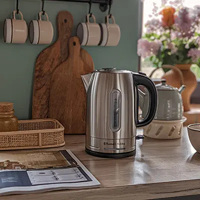 Na blacie kuchennym stoi czajnik ze stali nierdzewnej. W tle widoczne deski do krojenia, kwiaty w wazonie i wiszące kubki.