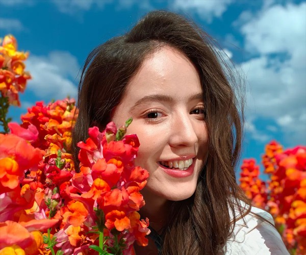 Portret w plenerze: osoba o długich brązowych włosach wśród gęstych pomarańczowo‑czerwonych kwiatów na tle błękitnego nieba z chmurami