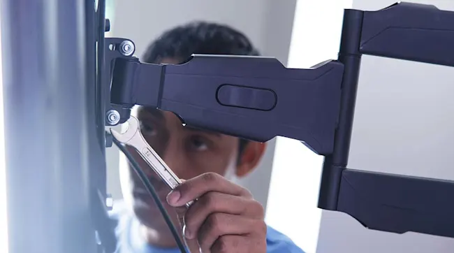 Osoba dokręca wspornik telewizora kluczem. Czarny wspornik jest przymocowany do metalowej rury.