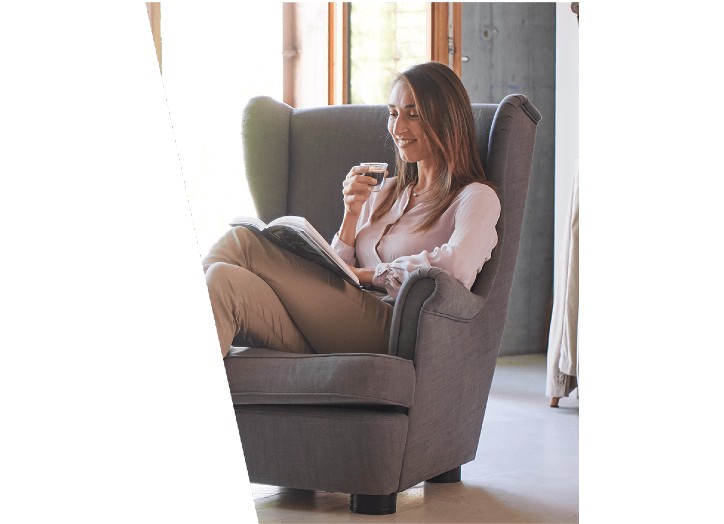 Kobieta siedzi w fotelu, trzymając filiżankę i czytając gazetę. Za nią widać okno i zasłony.