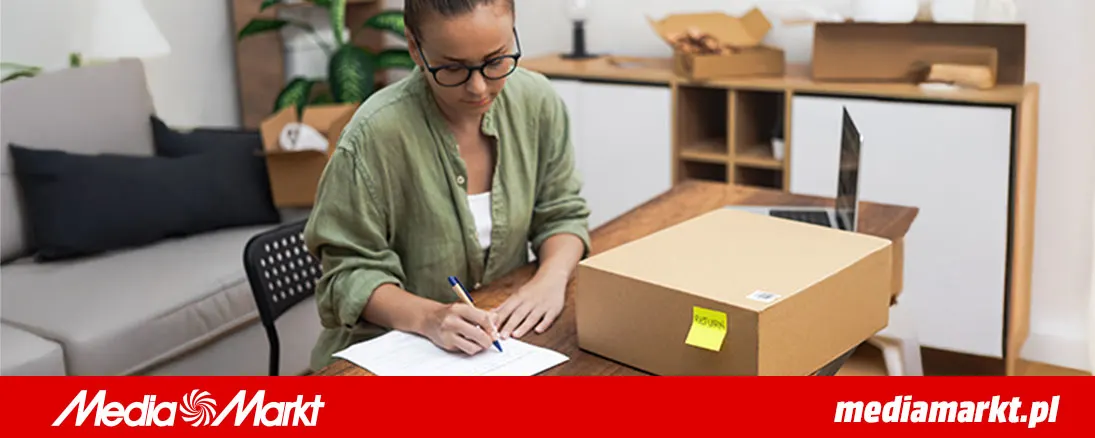 Kobieta w okularach pisze na kartce przy kartonie. W tle sofa, szafka i rośliny doniczkowe.