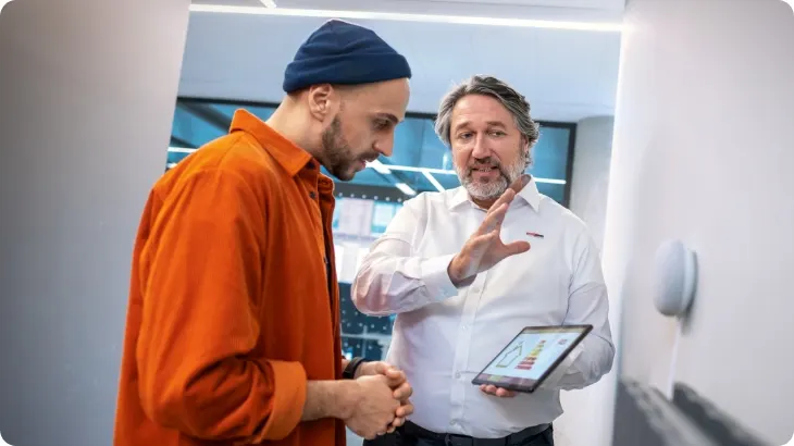 Na zdjęciu dwóch mężczyzn. Jeden w niebieskiej czapce i pomarańczowej koszuli, drugi w białej koszuli trzyma tablet z wykresami.