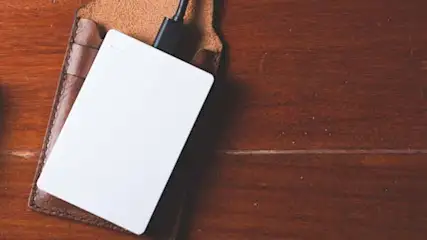 Na drewnianym stole leżą: laptop, telefon, biały dysk zewnętrzny podłączony kablem do brązowego etui.