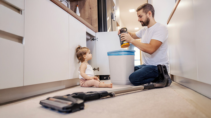 Mężczyzna wyrzuca śmieci, obok siedzi dziecko, na podłodze leży odkurzacz w jasnym wnętrzu.