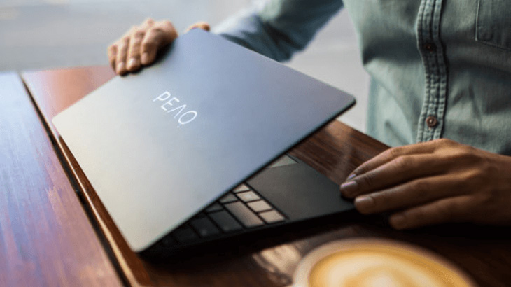 Osoba otwiera laptop na drewnianym stole. Widoczna klawiatura, napis PEAQ na obudowie i filiżanka kawy.