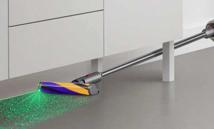 Odkurzacz z fioletowo-żółtą szczotką i zielonym laserem na szarej powierzchni. W tle białe szafki i podpora.