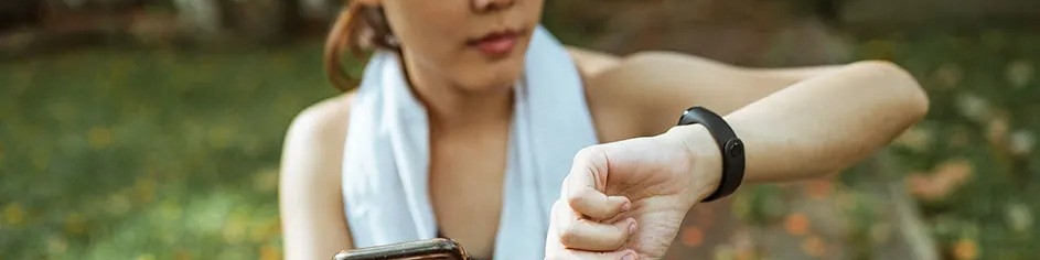 Kobieta w sportowym stroju sprawdza czarny smartwatch, trzymając telefon. Na szyi ma biały ręcznik, tło to zieleń.