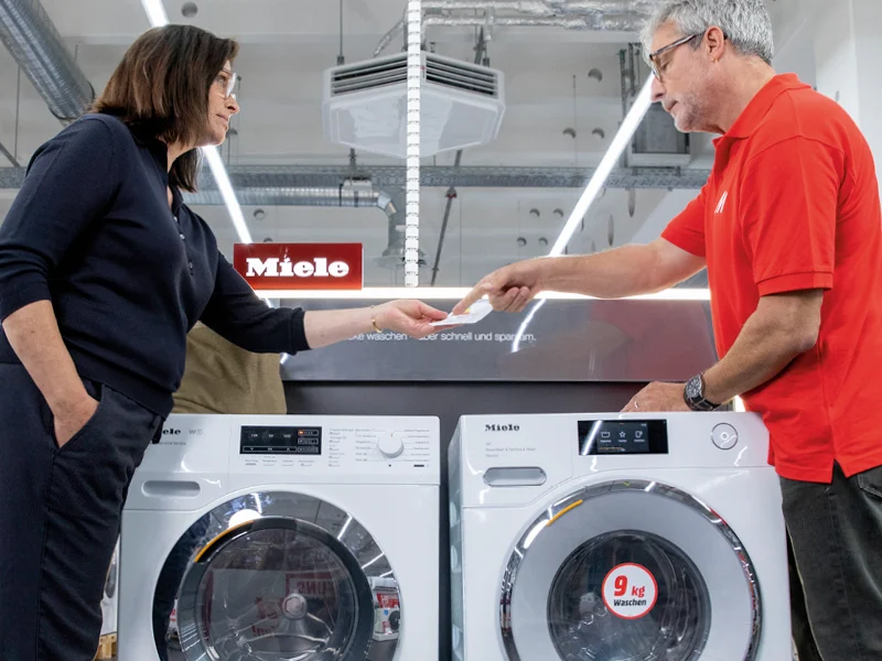 Kobieta i mężczyzna stoją przed dwiema białymi pralkami Miele, wymieniając się kartką w sklepie z oświetleniem sufitowym.