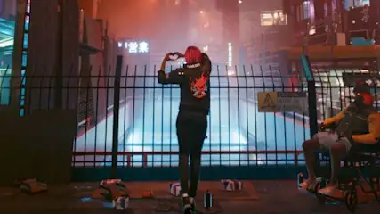 Na pierwszym planie metalowe ogrodzenie, za nim postać w czarnym ubraniu tworzy dłońmi kształt serca. Po prawej osoba na wózku.