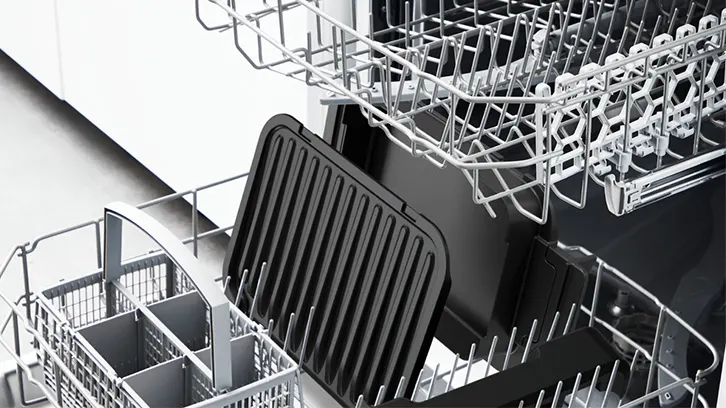 Wnętrze zmywarki z metalowymi koszami. Na dolnym koszu leży czarna patelnia grillowa i tacka.