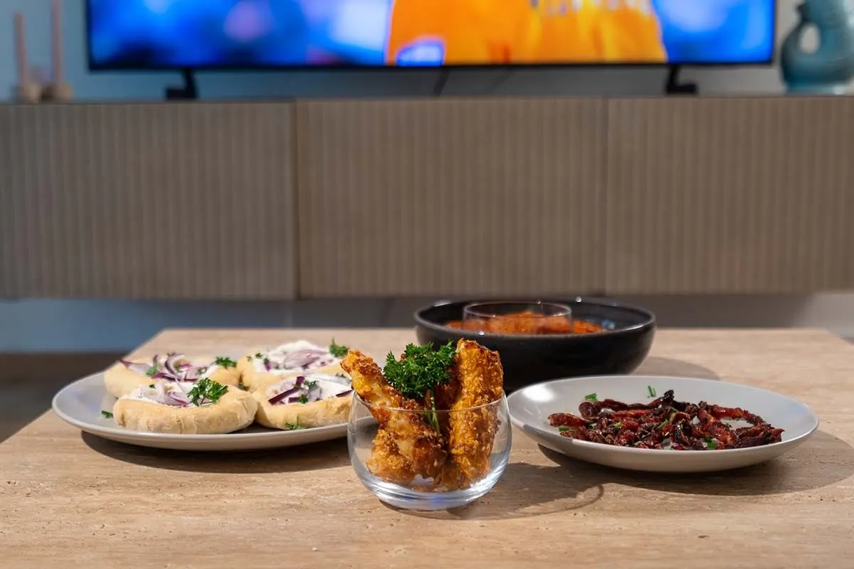 Différents finger foods sur une table devant la télévision, dont des petits pains garnis, des sticks croustillants et des snacks au lard. 