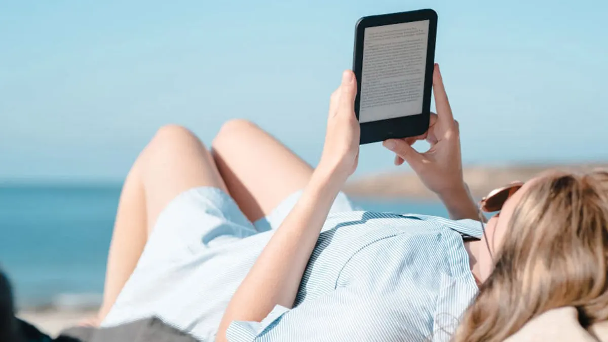 Eine Person liegt am Strand und liest auf einem E-Reader, mit blauem Himmel und Meer im Hintergrund.