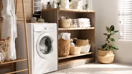 Eine weisse Waschmaschine steht in einem hellen Raum mit Holzregalen, in den regalen befinden sich Wäschekörbe und Wäsche
