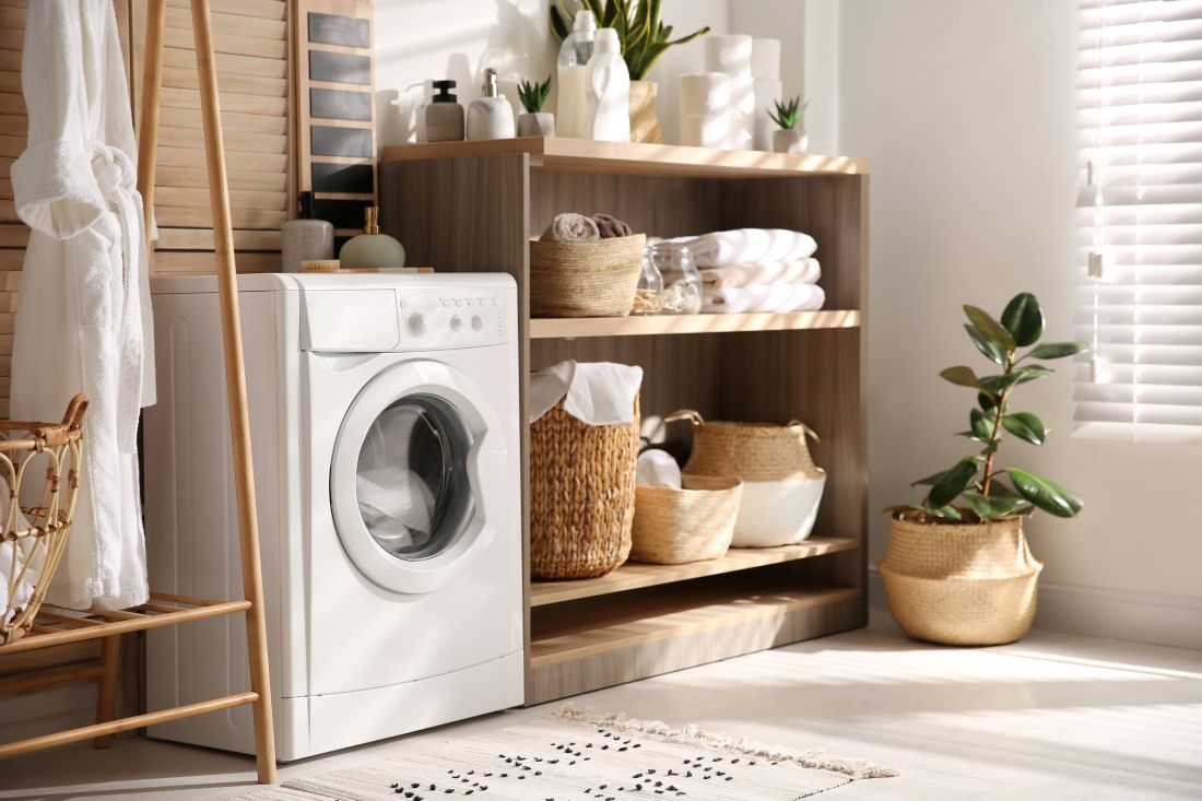 Una lavatrice bianca si trova in una stanza luminosa con scaffali in legno, sugli scaffali si trovano cestini per il bucato e della biancheria.