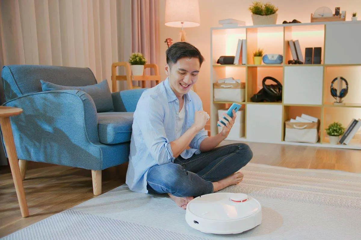 Uomo seduto sul pavimento del soggiorno con lo smartphone mentre un robot aspirapolvere bianco pulisce il tappeto.