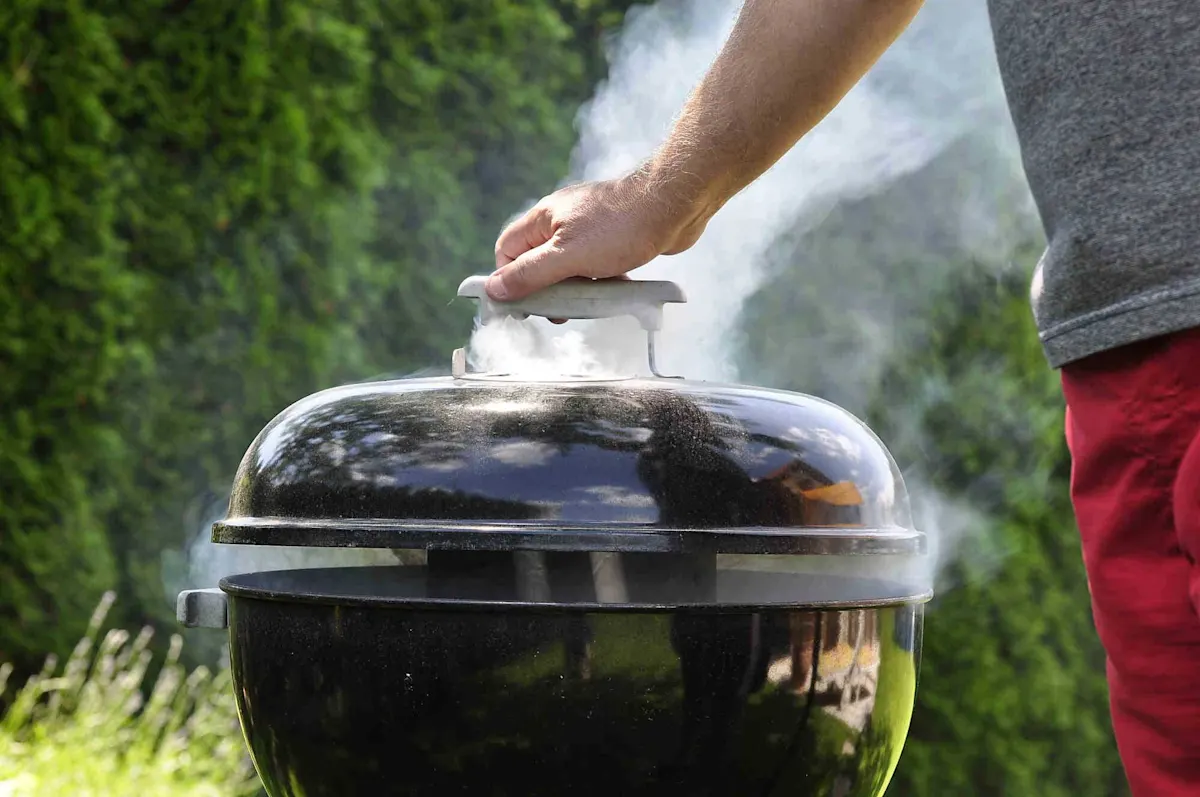 Persona che solleva il coperchio di un barbecue a sfera nero, mentre un denso fumo sale nel giardino. 
