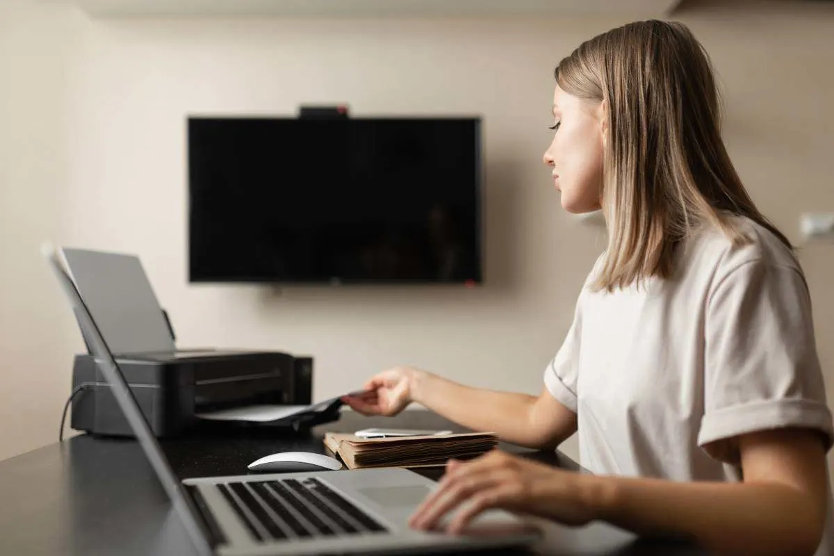 Donna che lavora al laptop e prende un foglio stampato dalla stampante. 