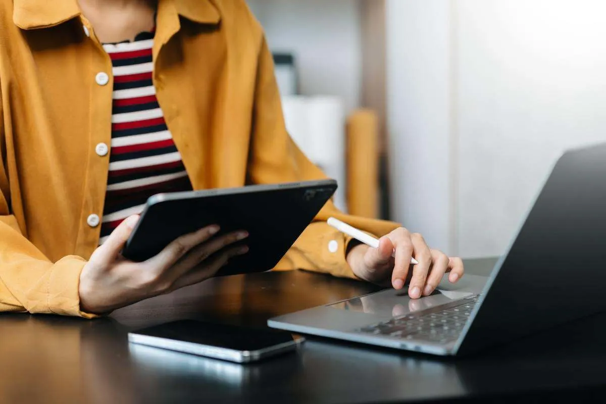 Une personne travaille à un bureau avec une tablette, un stylet, un ordinateur portable et un smartphone.