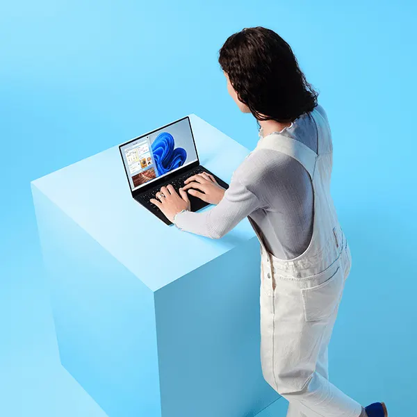 Eine hellgekleidete Frau tippt auf einem schwarzen Windows-Laptop, der auf einem hellblauen Podest steht.