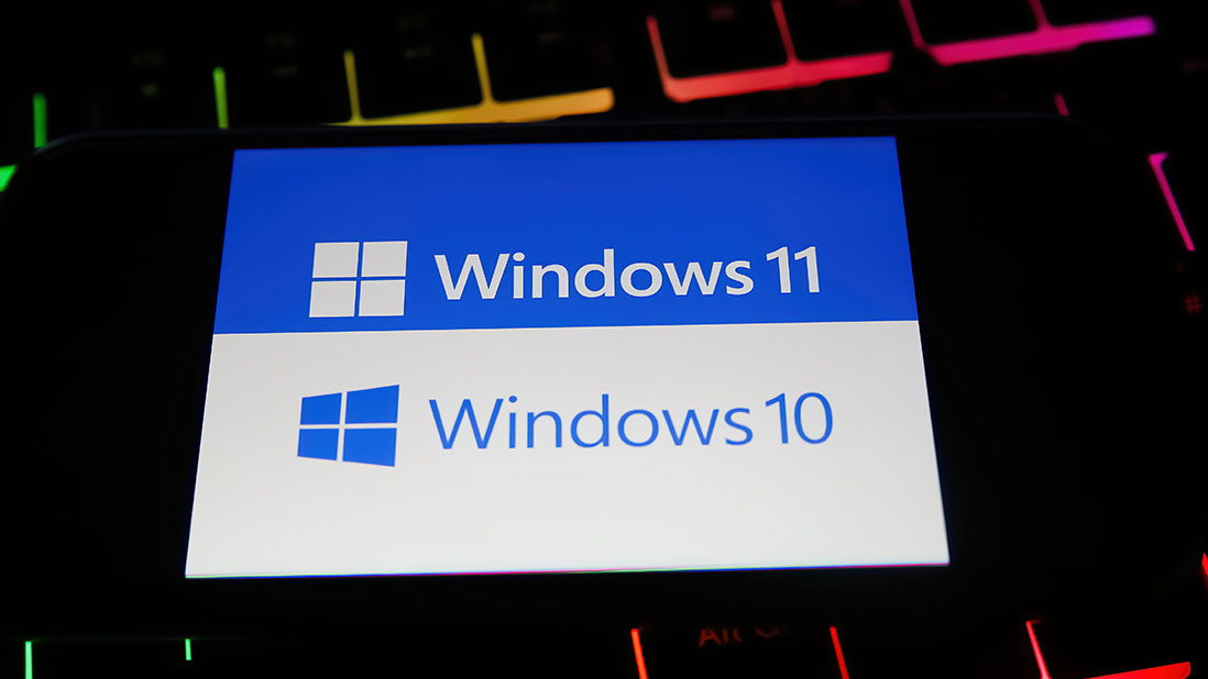 Schermo di smartphone che mostra loghi e testo "Windows 11" su sfondo blu e "Windows 10" su sfondo bianco, su tastiera retroilluminata.