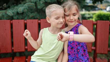 Zwei Kinder  sitzen auf einer Bank im freien und schauen auf die TCL-Kinder-Smartwatch auf dem Handgelenk des Mädchens.