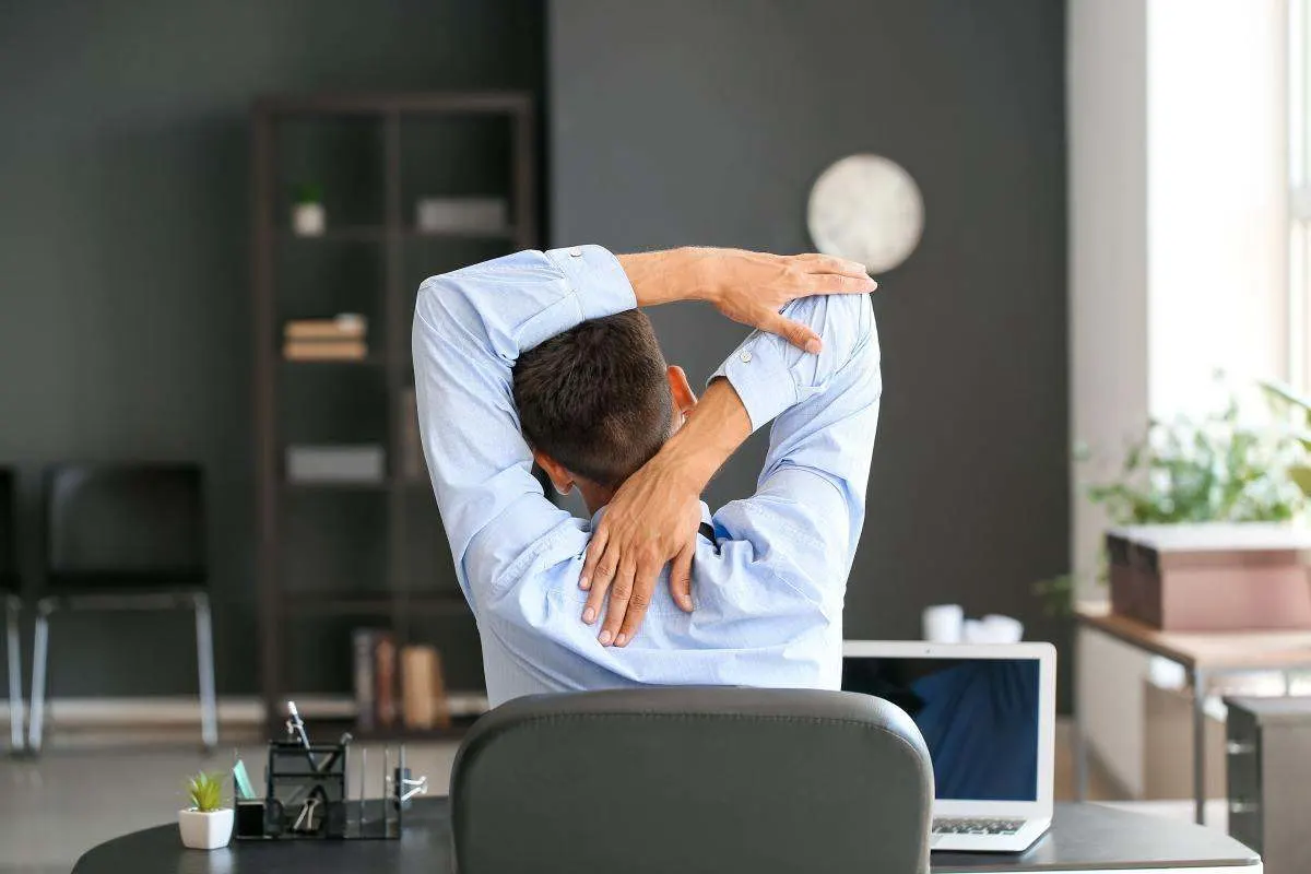 Mann sitzt am Schreibtisch vor einem Laptop und dehnt mit erhobenen Armen Schultern und oberen Rücken.