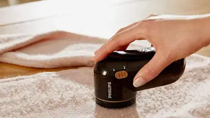 Hand hält schwarzen Philips Fusselrasierer über einem hellrosafarbenen Frotteehandtuch auf einer Holzoberfläche.