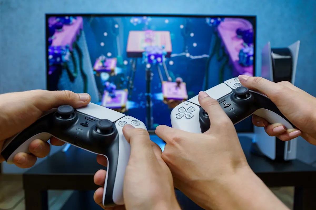 Deux personnes jouent sur une PlayStation 5 : gros plan de deux manettes DualSense devant un téléviseur avec un jeu coopératif coloré ; la console est à droite.