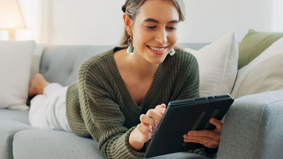 Eine Frau liegt auf einer grauen Couch und benutzt ein schwarzes Tablet. Kissen sind im Hintergrund sichtbar.