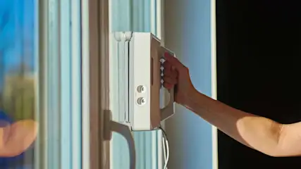 Hand hält einen weissen Fensterputzroboter, der an einer Fensterscheibe angebracht ist.